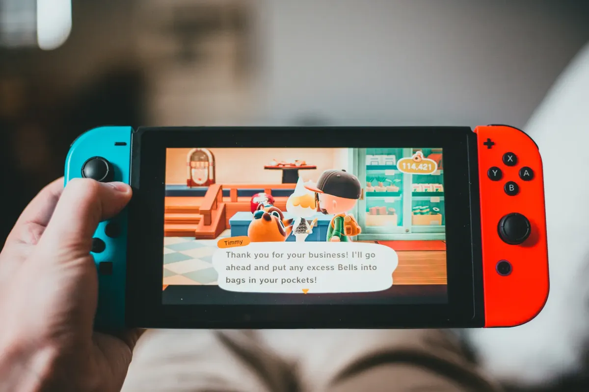 クロップされた匿名な人物がゲームコンソールを使ってゲームをプレイしている。背景にはぼかしたホームのシーンがあります。