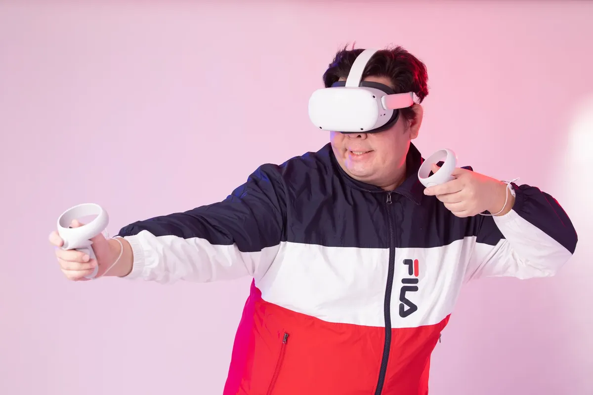 Adult man wearing VR headset and using controllers, immersed in a virtual reality game against a pink studio backdrop.